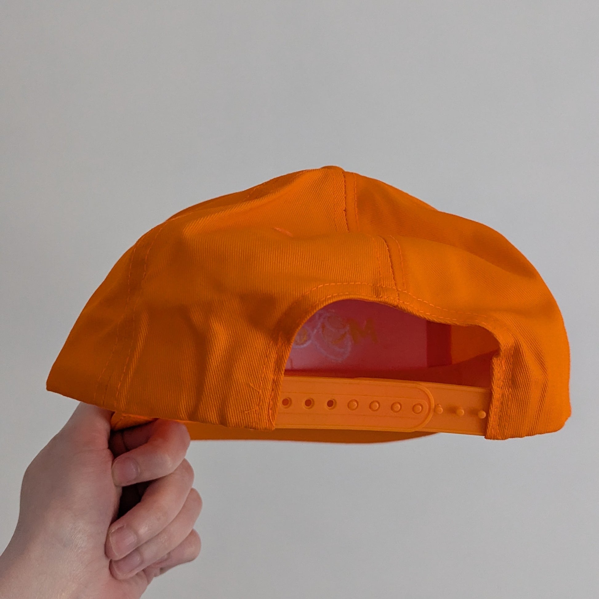 Orange cap held by a hand, showing snap adjuster, against a plain background