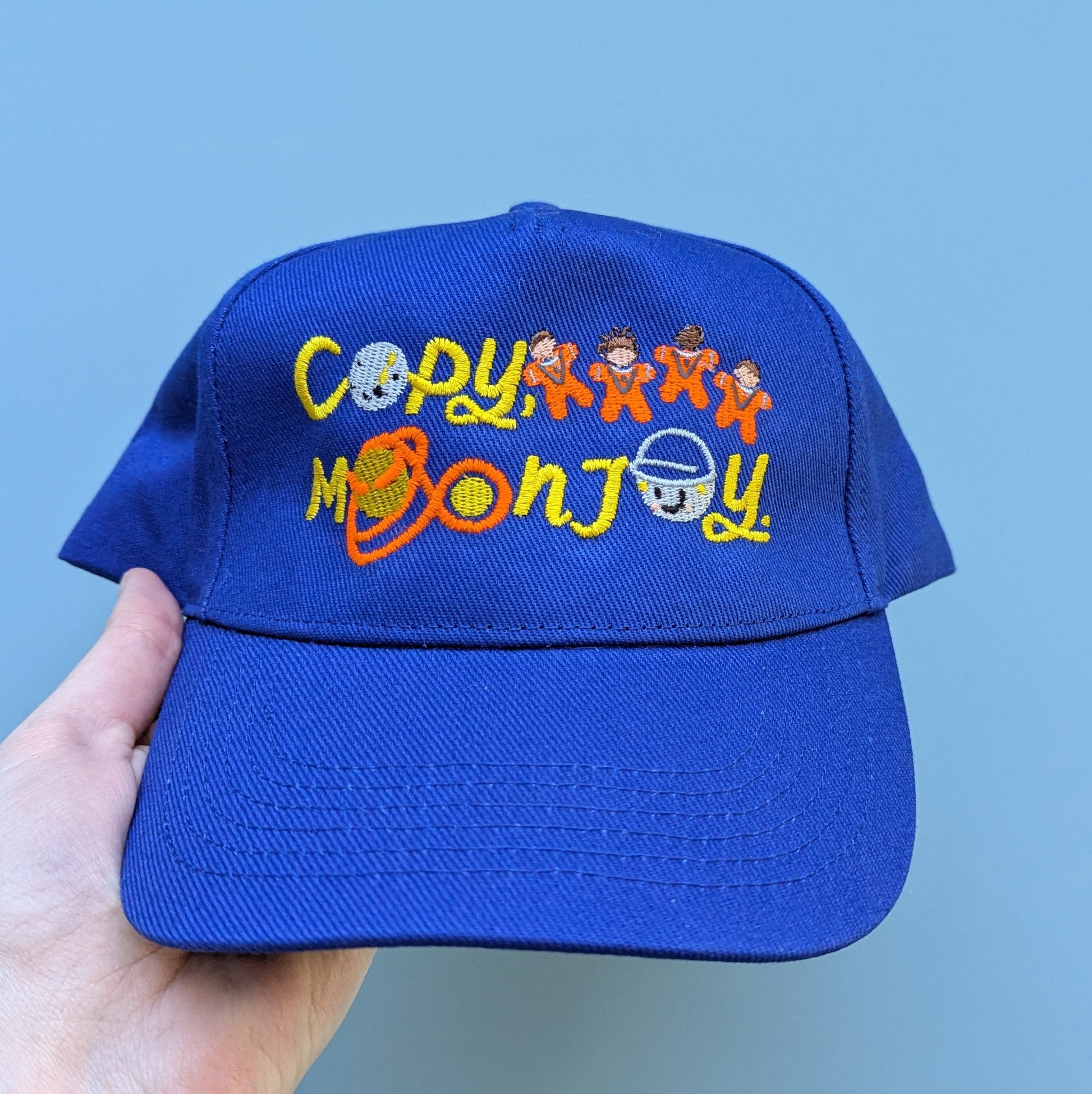 Bright blue adults blue curved cap with colourful text that reads Copy, Moon Joy. Accompanying text includes graphics.The product is held by a hand against a light blue background.