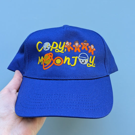 Bright blue adults blue curved cap with colourful text that reads Copy, Moon Joy. Accompanying text includes graphics.The product is held by a hand against a light blue background.
