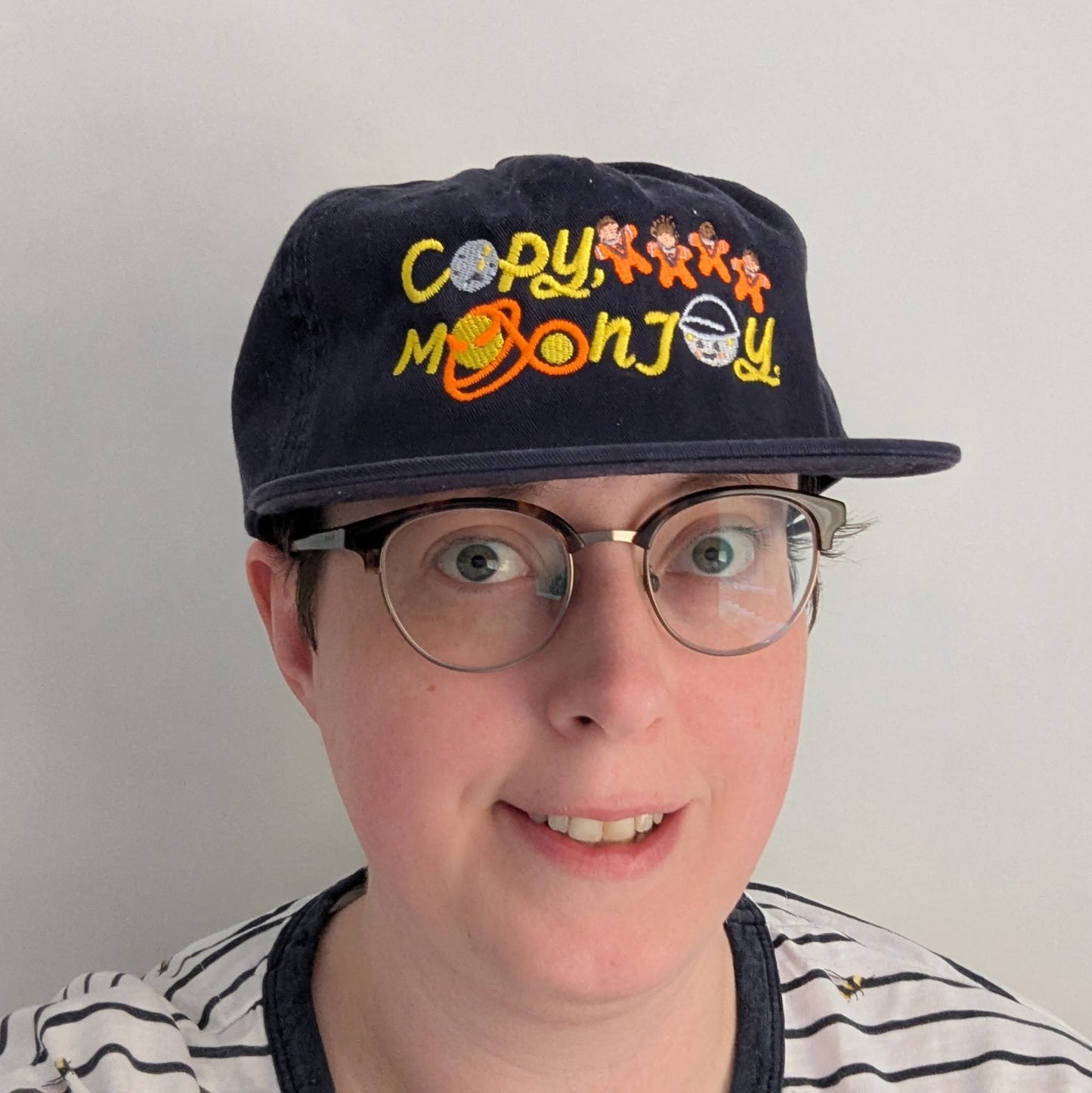 A photo showing a front faced person wearing a navy blue cap with colourful text that reads Copy, Moon Joy. 

Accompanying text includes graphics.The product is held by a hand against a white background.