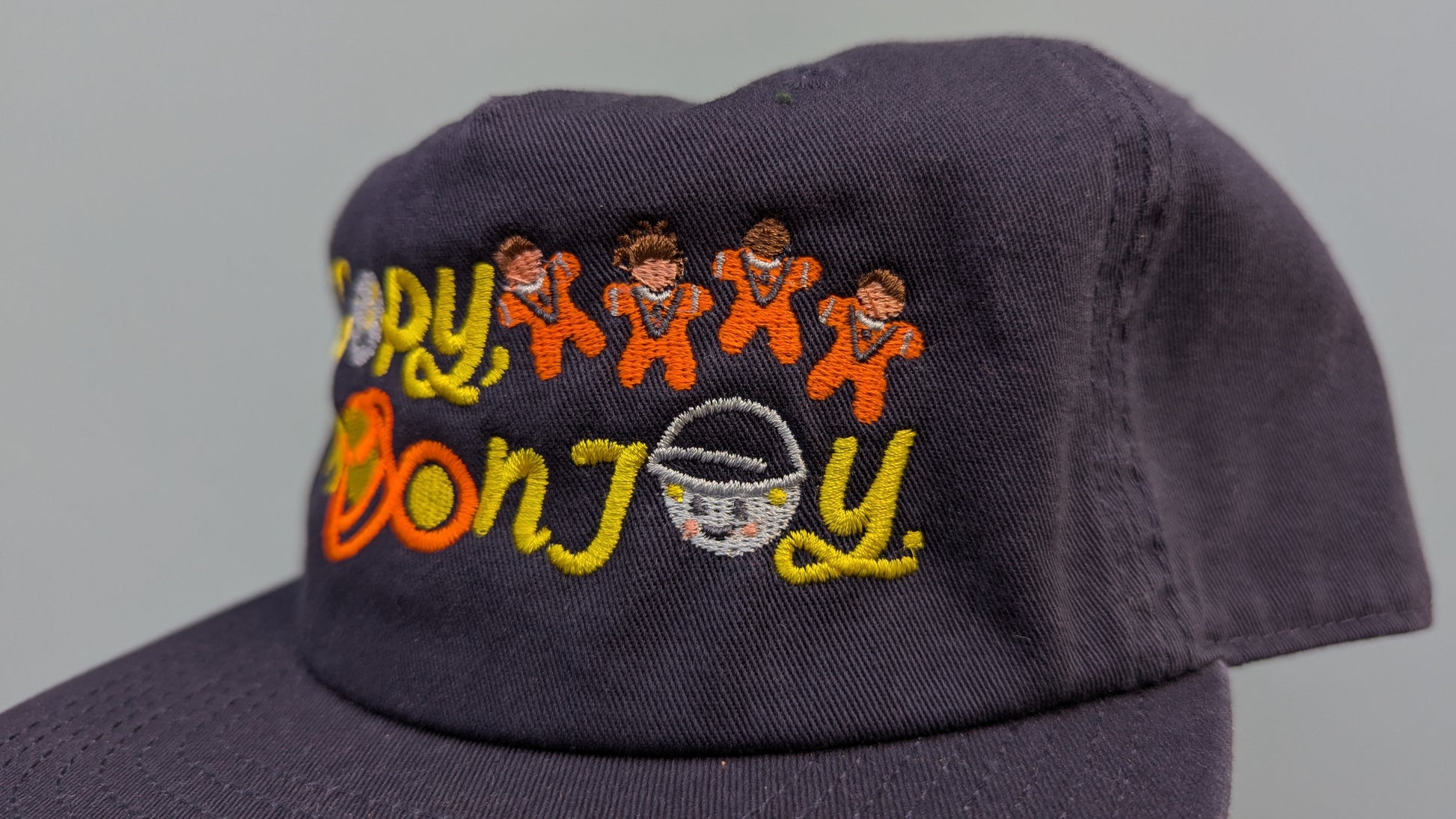 Navy blue cap with colourful text that reads Joy. Accompanying text includes graphics of the Artemis II crew and Rise the Gravity Indicator. The product is held by a hand against a light blue background.