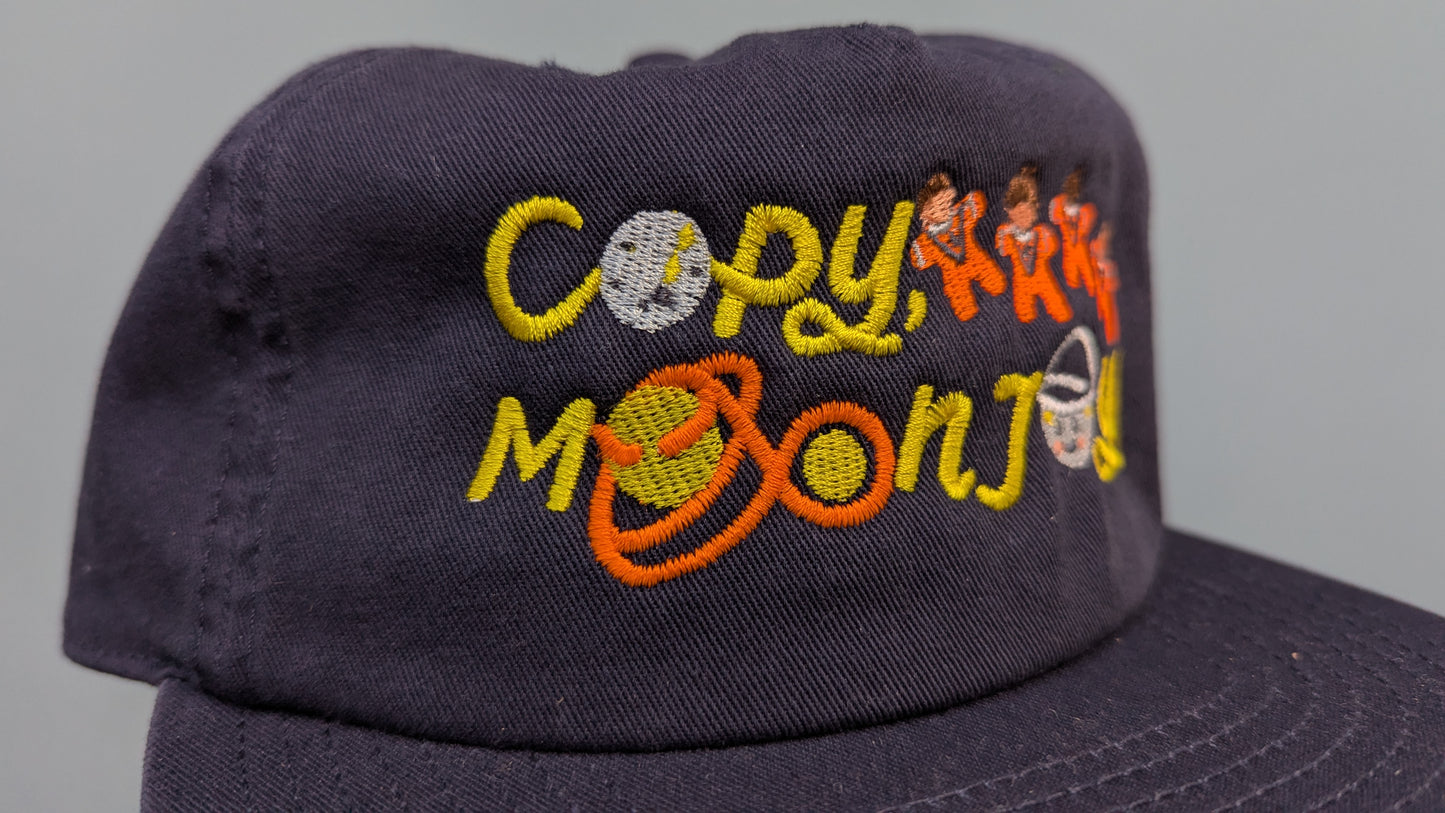 Navy blue cap with colourful text that reads Copy, Moon. Accompanying text includes graphics of the route Artemis II took and Moon with two craters after Carroll and Integrity.The product is held by a hand against a light blue background.