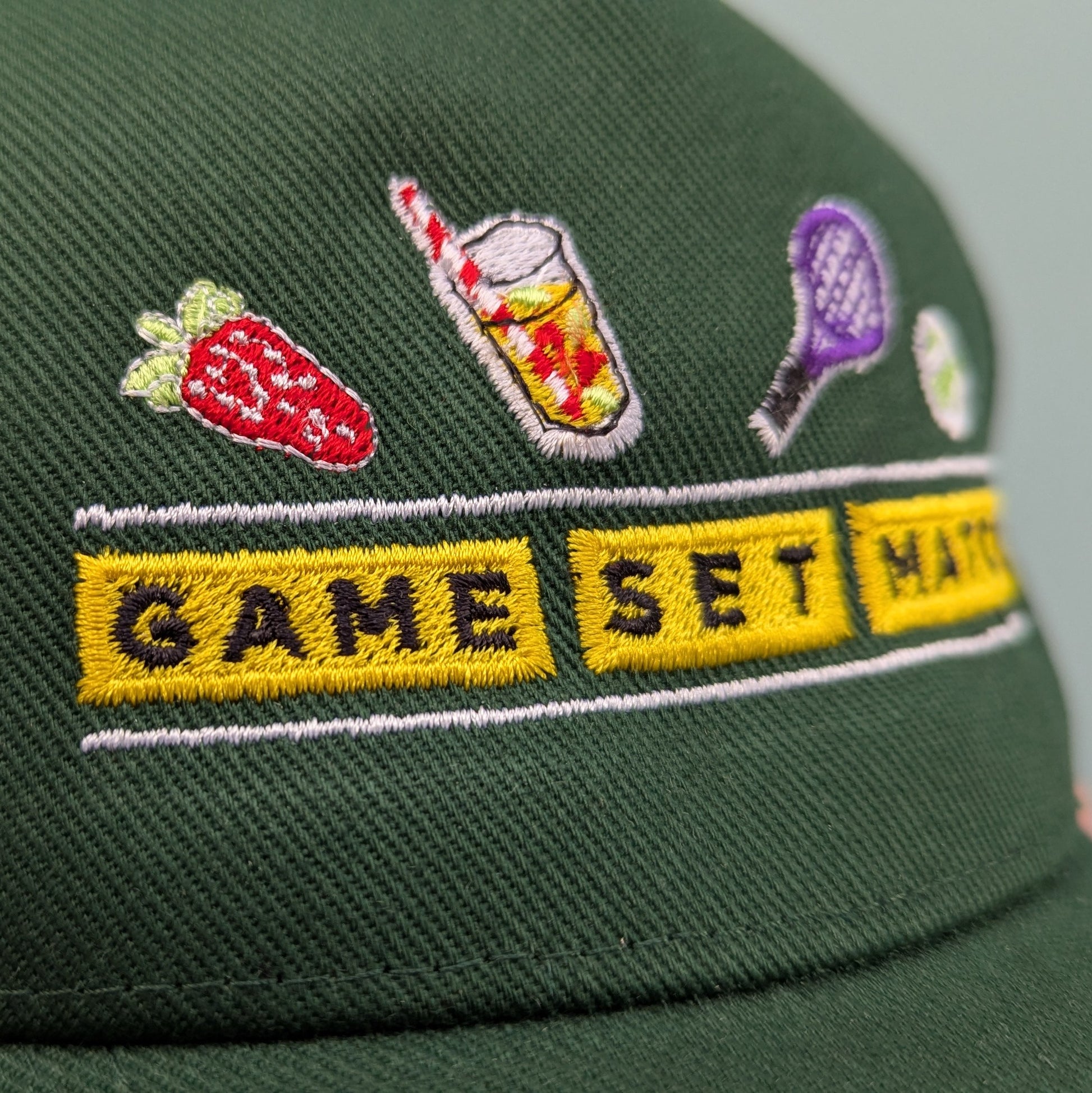 Bottle green adults curved cap with colourful text that reads Game Set Match. Accompanying text includes a close up of the graphics depicting a strawberry and a a drink with a red and white straw. The product is held against a light blue background.