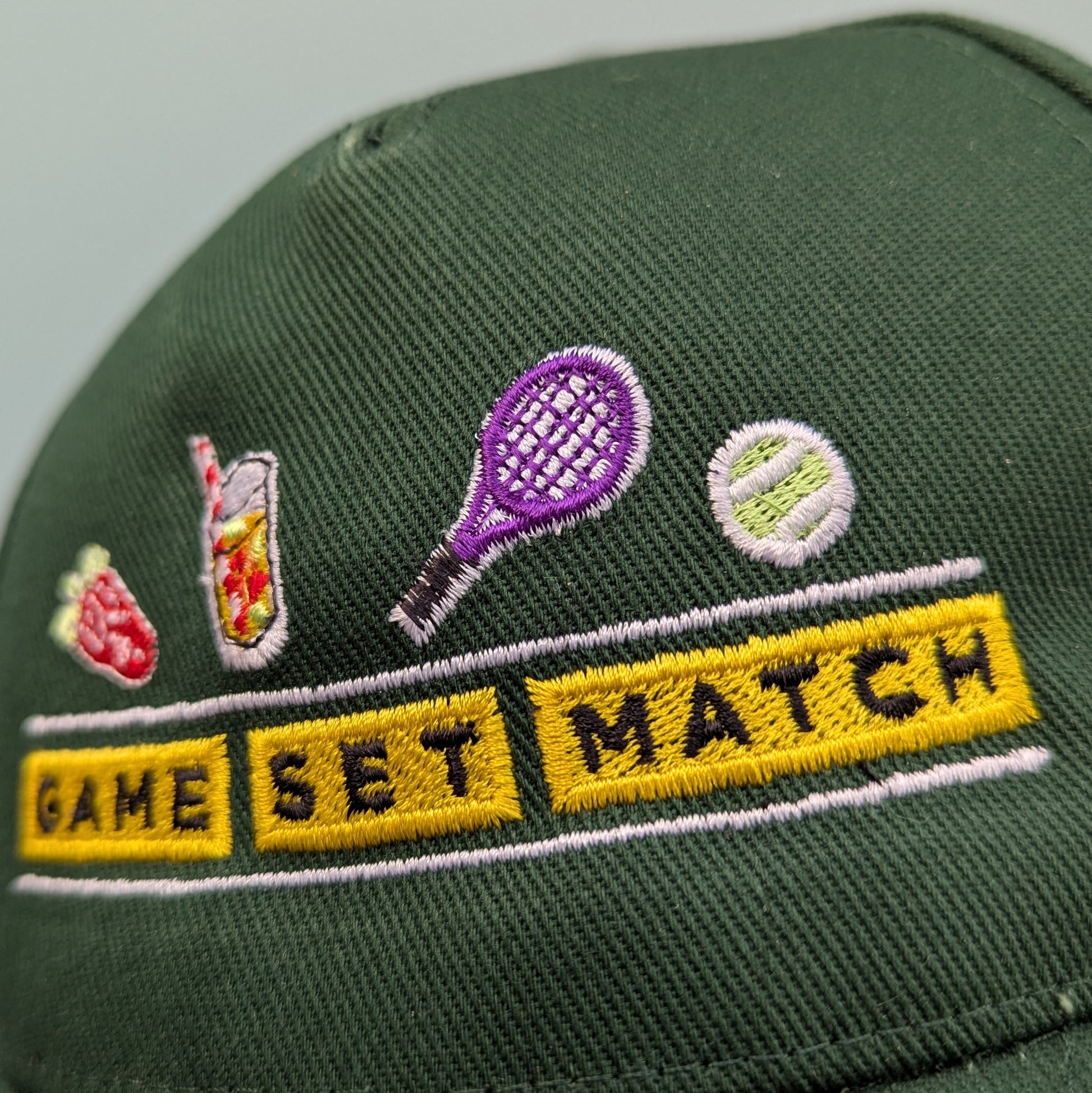 Bottle green adults curved cap with colourful text that reads Game Set Match. Accompanying text includes a close up of the graphics depicting a tennis racket and a tennis ball The product is held against a light blue background.