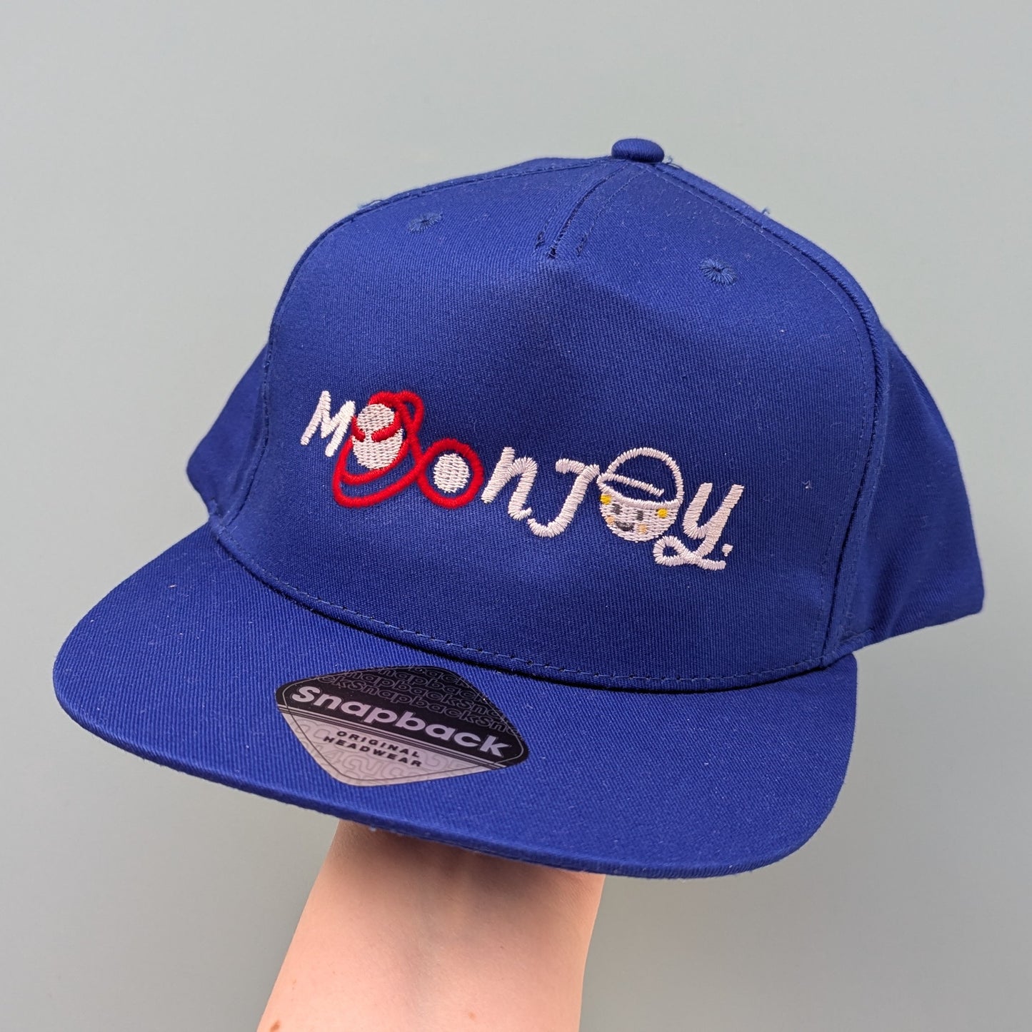 Blue snapback cap with 'Moon Joy' text, featuring a red route and Rise, held by a hand against a light background.