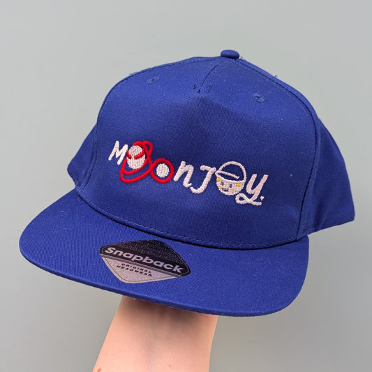 Blue snapback cap with 'Moon Joy' text, featuring a red route and Rise, held by a hand against a light background.