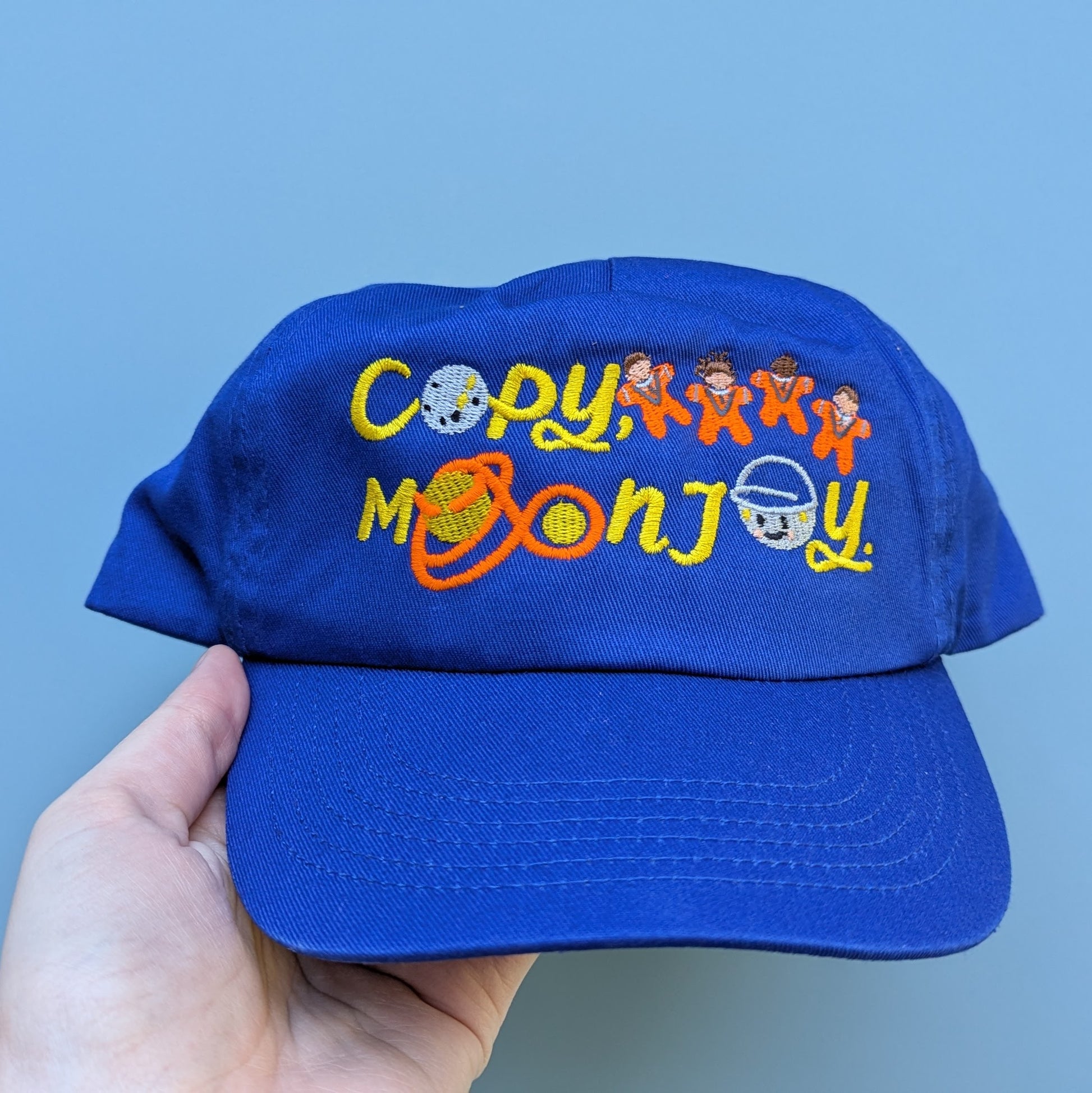Bright blue kids blue curved cap with colourful text that reads Copy, Moon Joy. Accompanying text includes graphics.The product is held by a hand against a light blue background.