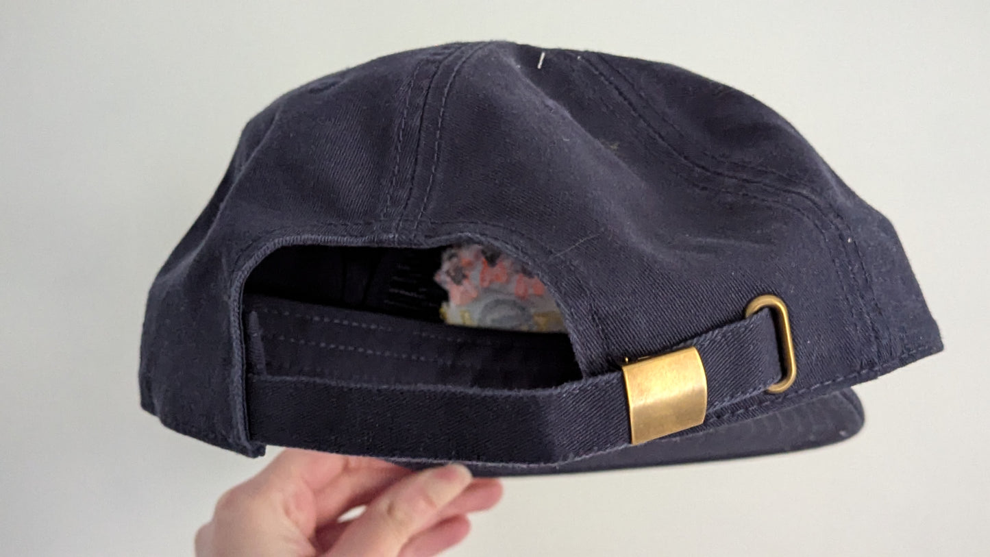 Image shows a goldish metal clasp fastener at the back of a navy cap, ensuring adjustability to the cap.