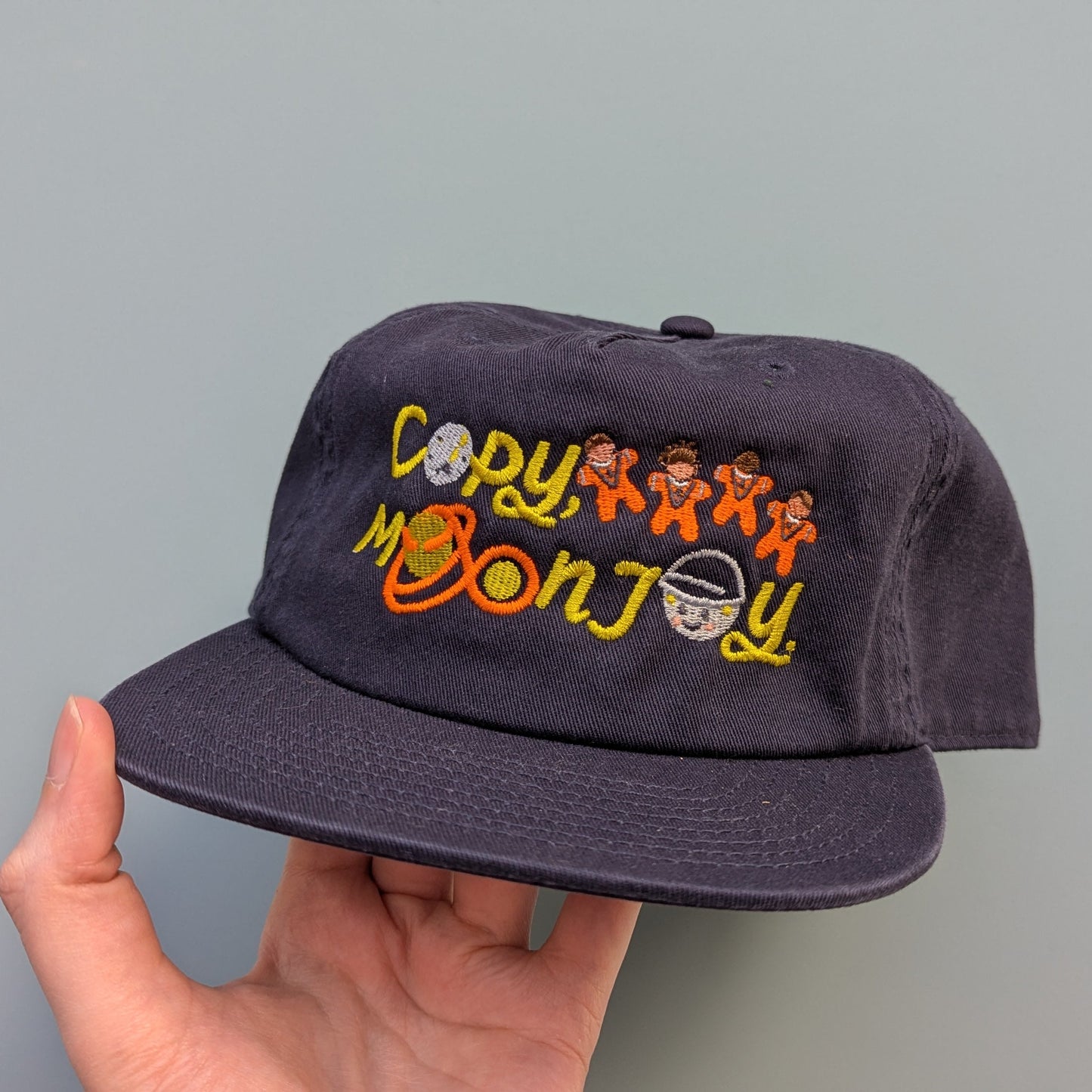 Navy blue cap with colorful text that reads Copy, Moon Joy. Accompanying text includes graphics.The product is held by a hand against a light blue background.