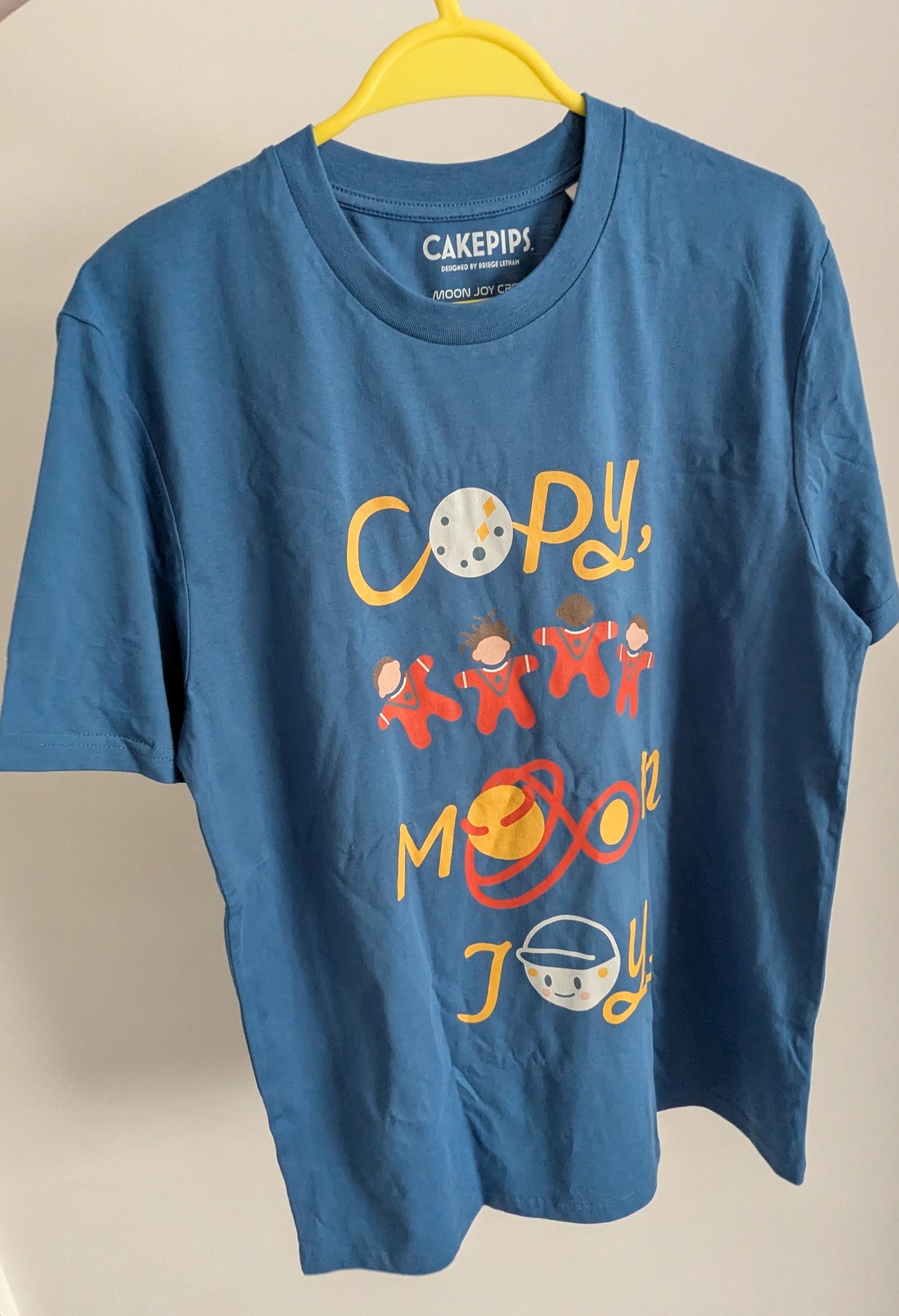 Mindful blue t-shirt with colourful graphics and text saying 'copy, moon joy' , displayed on a hanger.