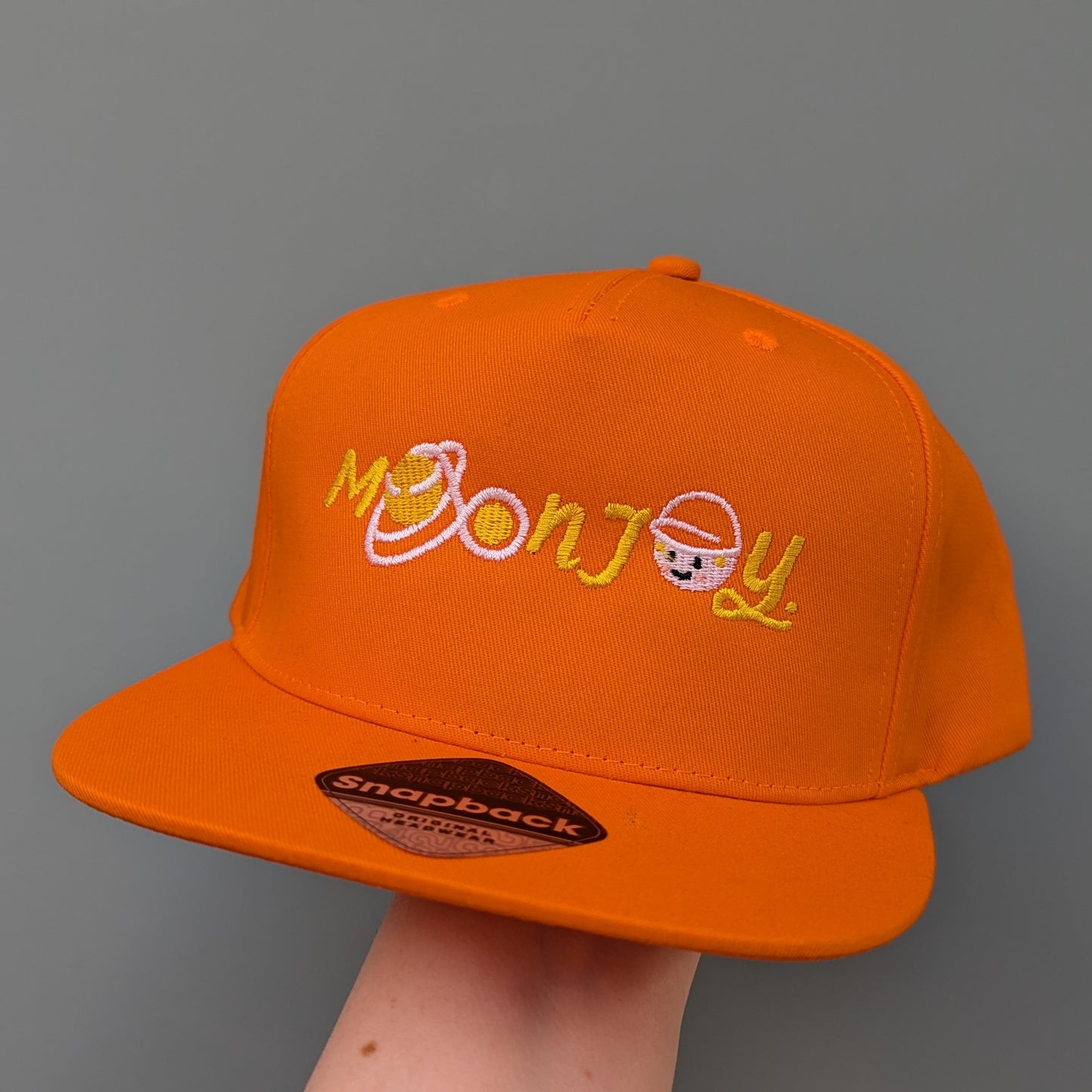 Orange snapback cap with 'Moon Joy' text, featuring a white route and Rise, held by a hand against a light background