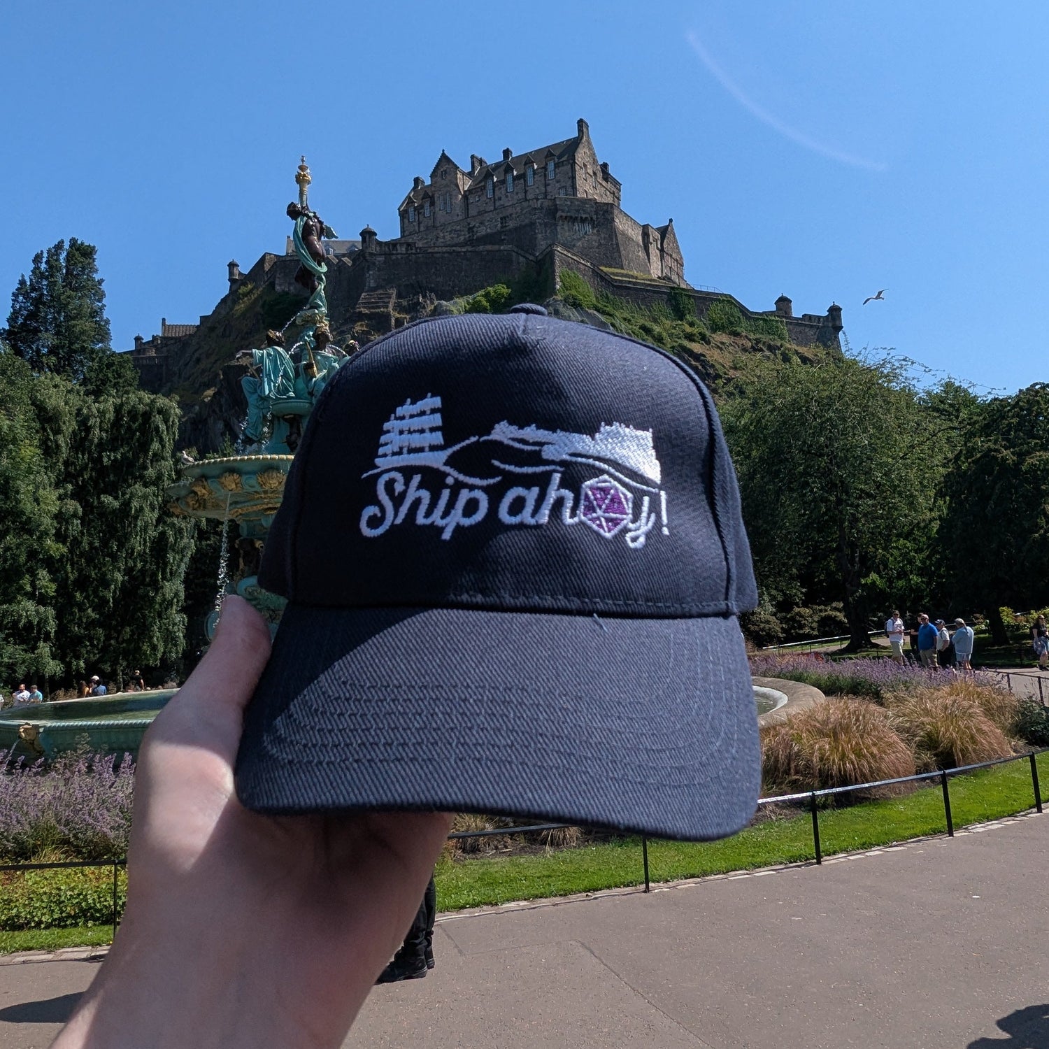 A navy blue cap with 'Ship ahoy!' embroidered on the front is held up by a hand against a background of Edinburgh Castle and a clear blue sky. It features a ship, Edinburgh Castle and a D20 dice with the date on it.
