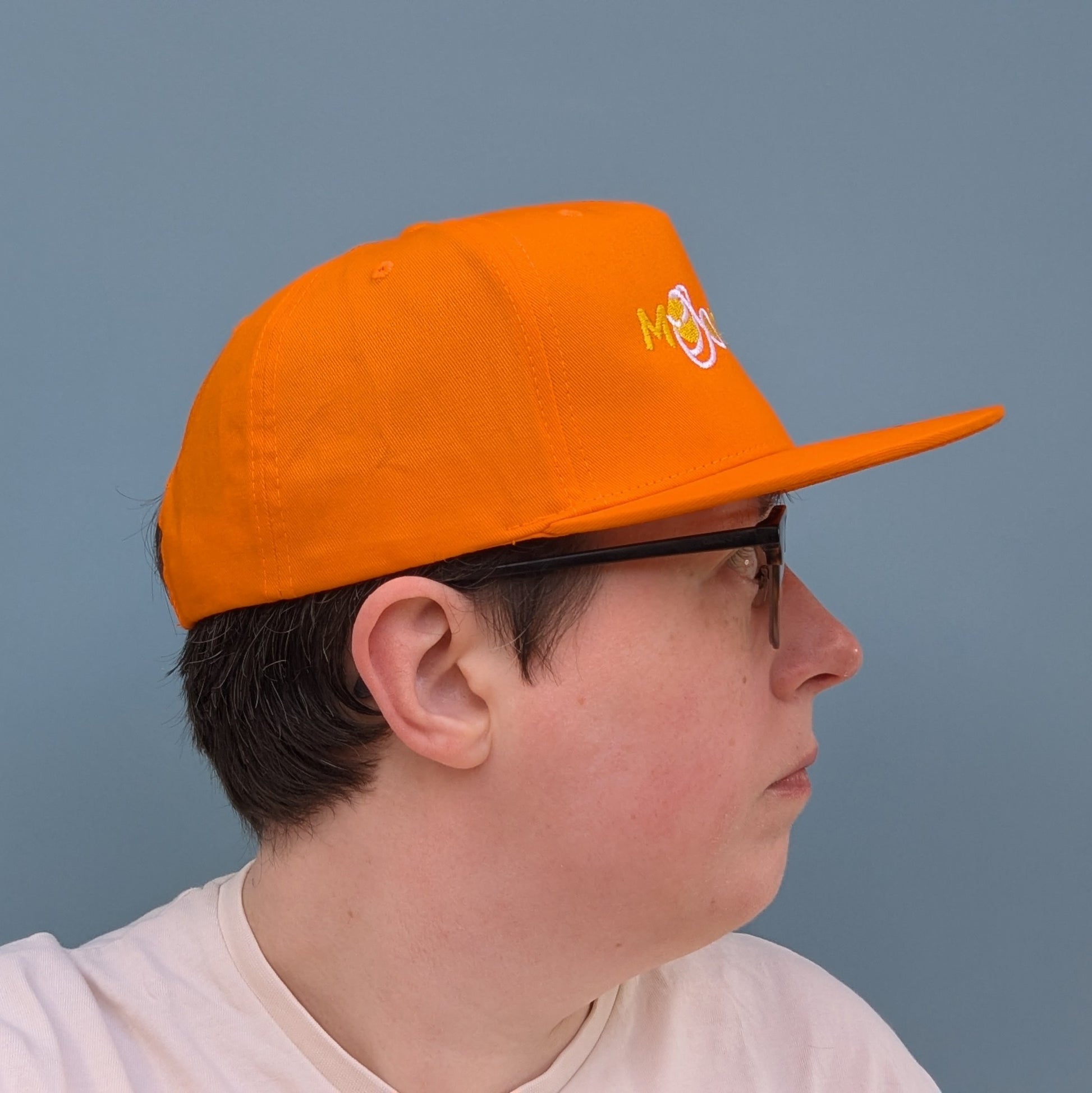 Side facing person wearing orange snapback cap with 'Moon Joy' text, featuring a white route and Rise, against a light background