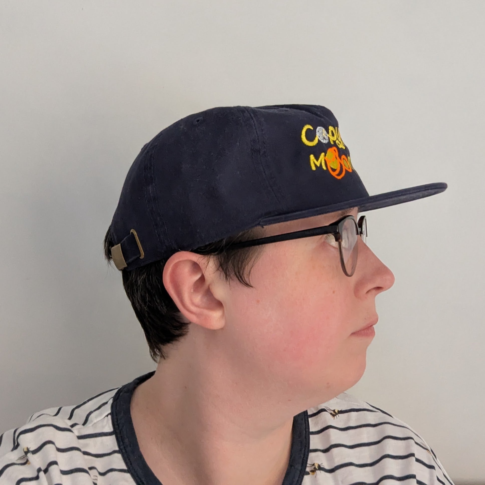 A photo showing a side of the face of person wearing a navy blue cap with colourful text that reads Copy, Moon Joy. 

Accompanying text includes graphics.The product is held by a hand against a white background.
